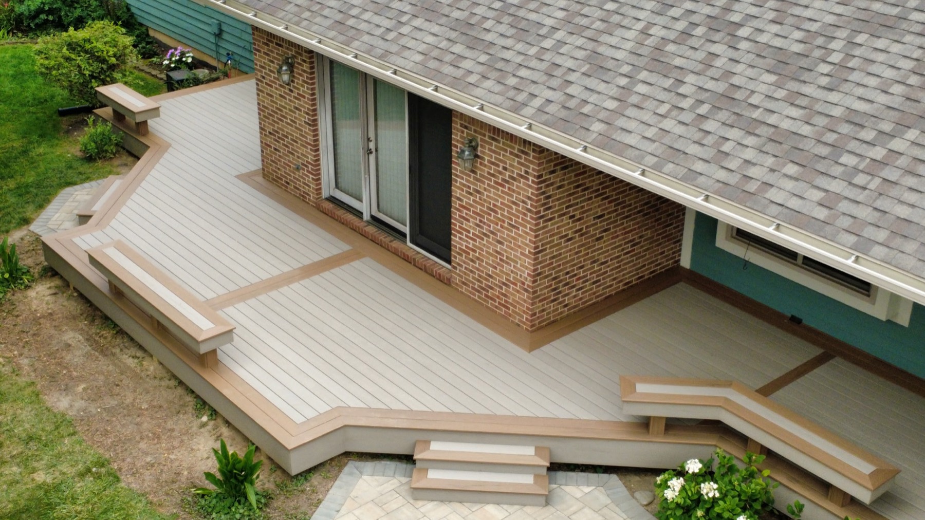 Aerial view of a light grey composite deck wrapping a brick home, with contrasting brown borders, two built-in benches and steps down to a paver patio