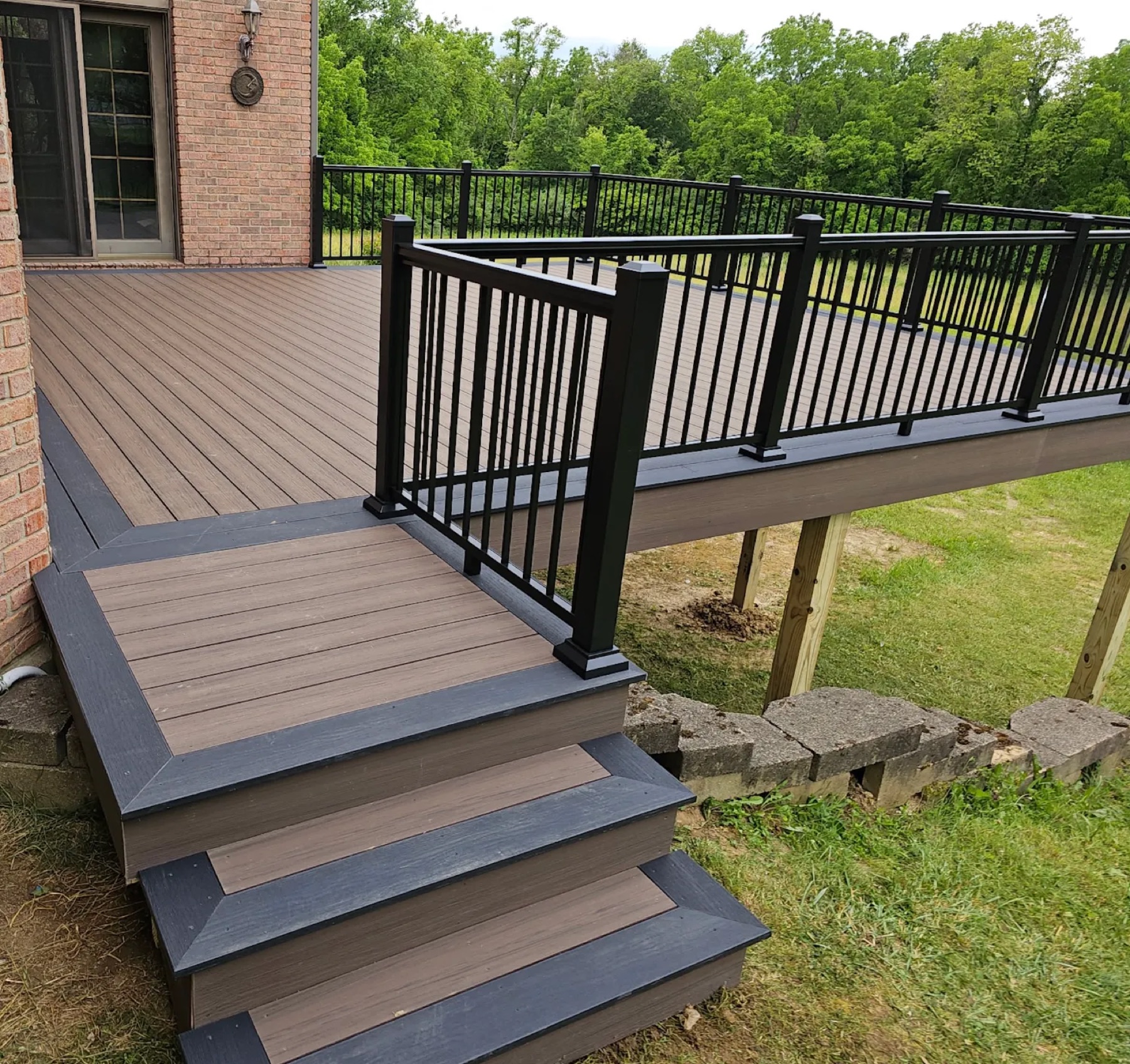 Brown composite deck with charcoal borders, black aluminum railing and a wide stair landing beside a brick home