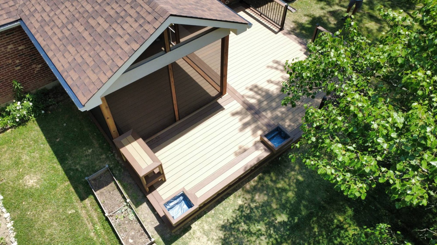 Aerial view of a tan composite deck with a gabled roof over one end, a built-in bench and two built-in planters