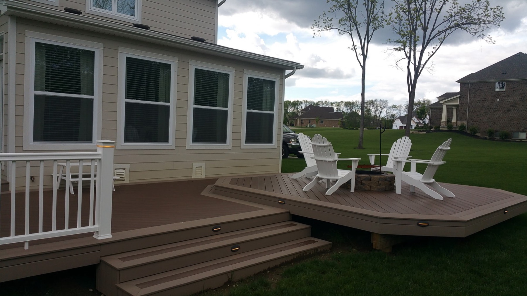 Lit composite steps leading from a main deck down to a fire-pit level with white Adirondack chairs