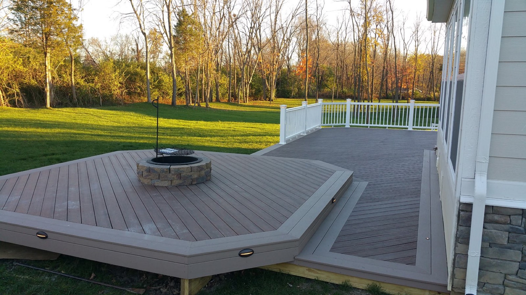 Angled lower deck level with a stone fire pit, edge lighting and white railing on the main deck, at sunset