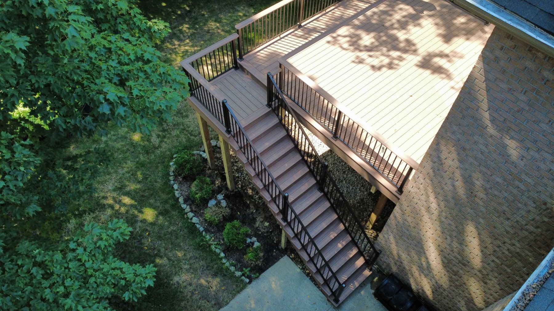 Aerial view of a raised brown composite deck on a brick home with black railing and a long stair run down to a concrete patio