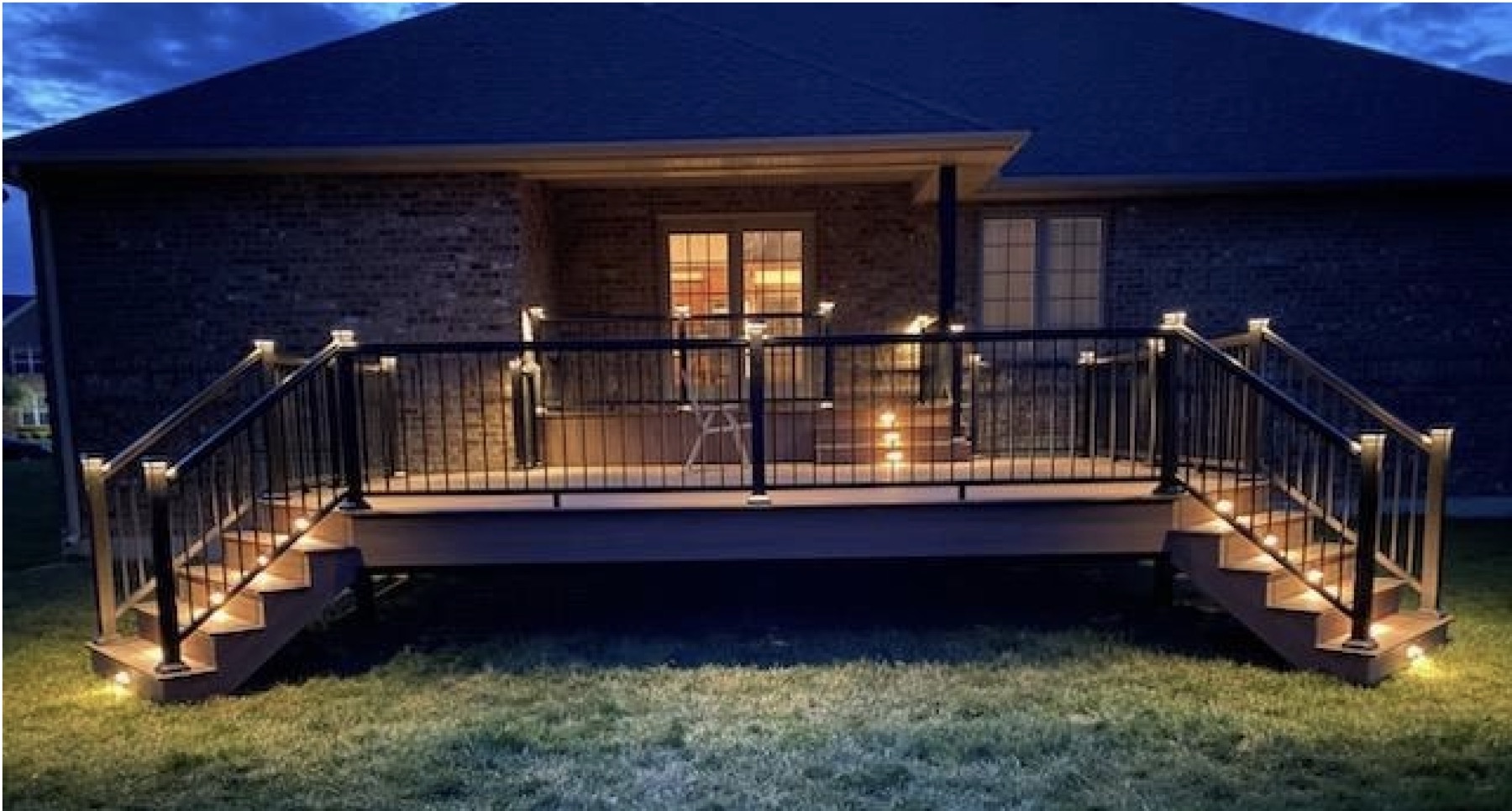 Composite deck at dusk with lit stair risers and glowing post caps on black aluminum railing