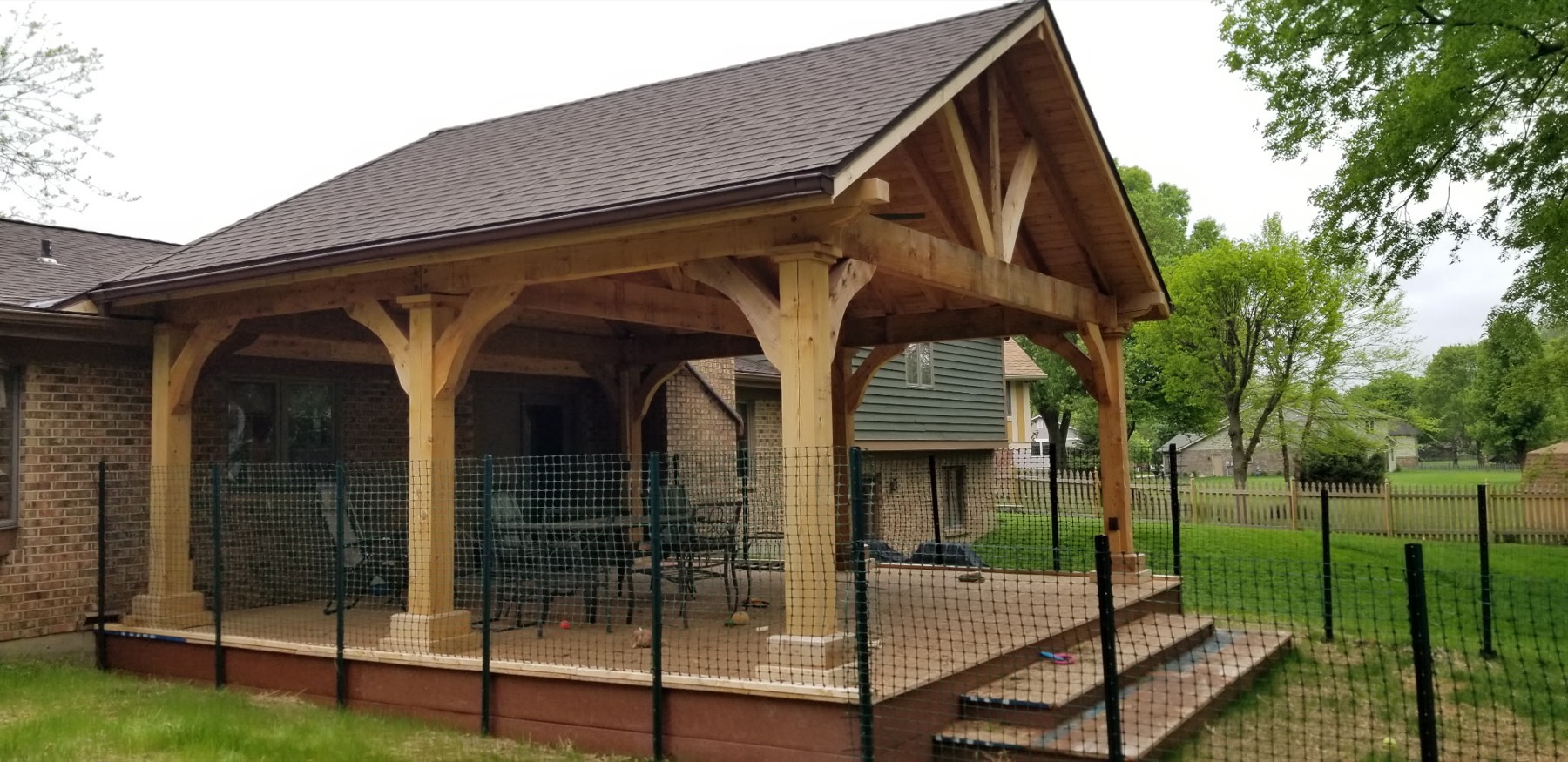 Timber-frame pavilion with a gabled shingle roof, exposed beams and knee braces over a composite deck behind a brick home