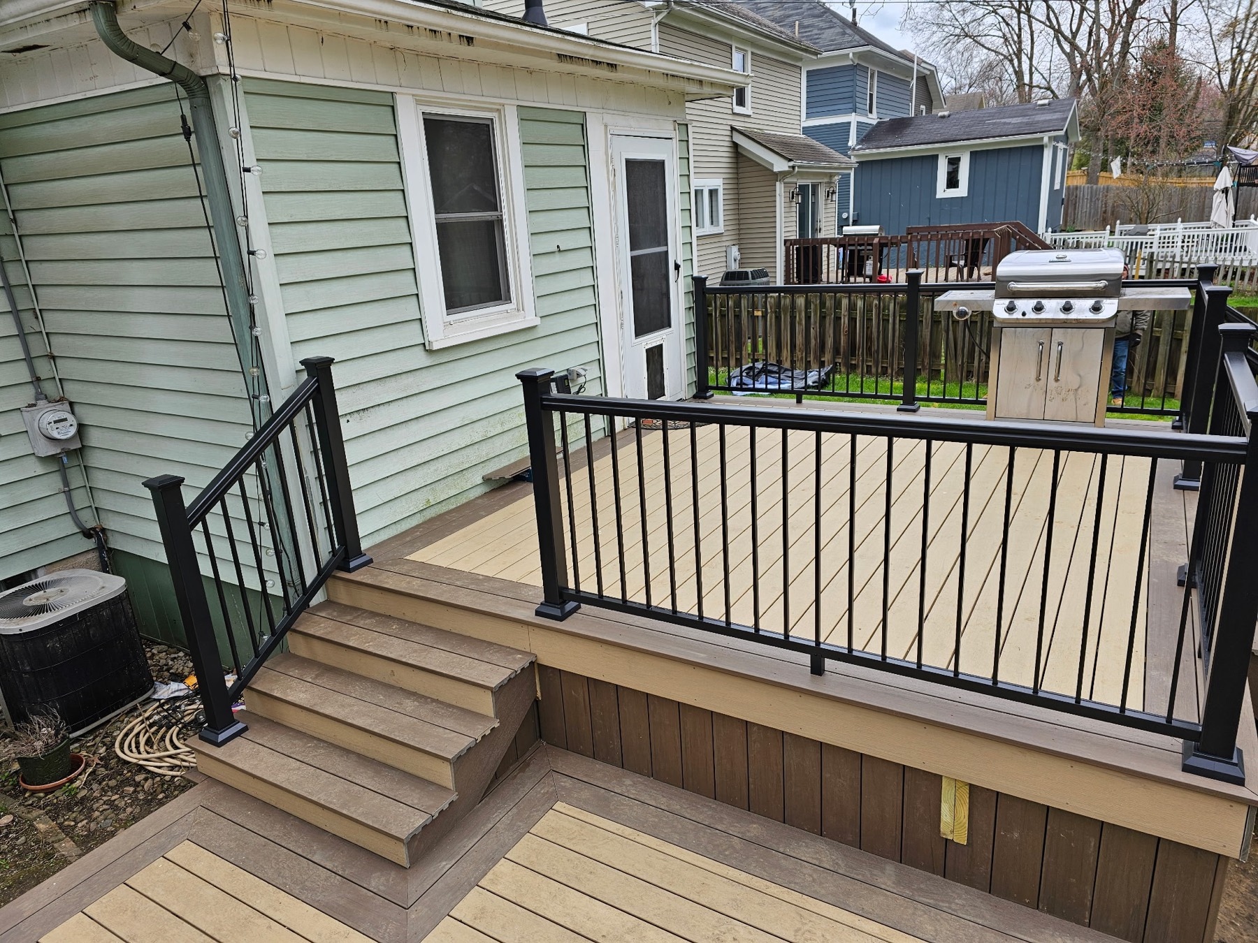 Two-level tan composite deck with dark brown borders and skirting, black railing and a grill